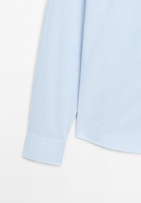 Manche longue d'une chemise bleu clair avec de fines rayures blanches verticales et un poignet boutonné sur fond blanc.