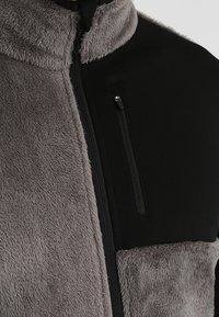 Närbild av en grå och svart fleecejacka med en vertikal dragkedja och en ficka med dragkedja på bröstet i den svarta sektionen.