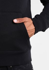 Felpa nera con tasca a marsupio. Realizzata in tessuto liscio e elasticizzato. Presenta polsini e orlo a coste. Mano posizionata nella tasca.