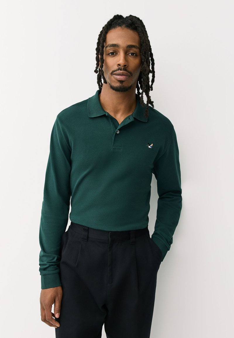 Hombre con rastas de longitud media lleva un polo de manga larga verde oscuro y pantalones negros, de pie con una mano en el bolsillo contra un fondo blanco.