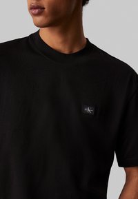 Czarna bawełniana koszulka z krótkim rękawem i okrągłym dekoltem, z prostokątną naszywką z logo CK po lewej stronie piersi. Gładka faktura.