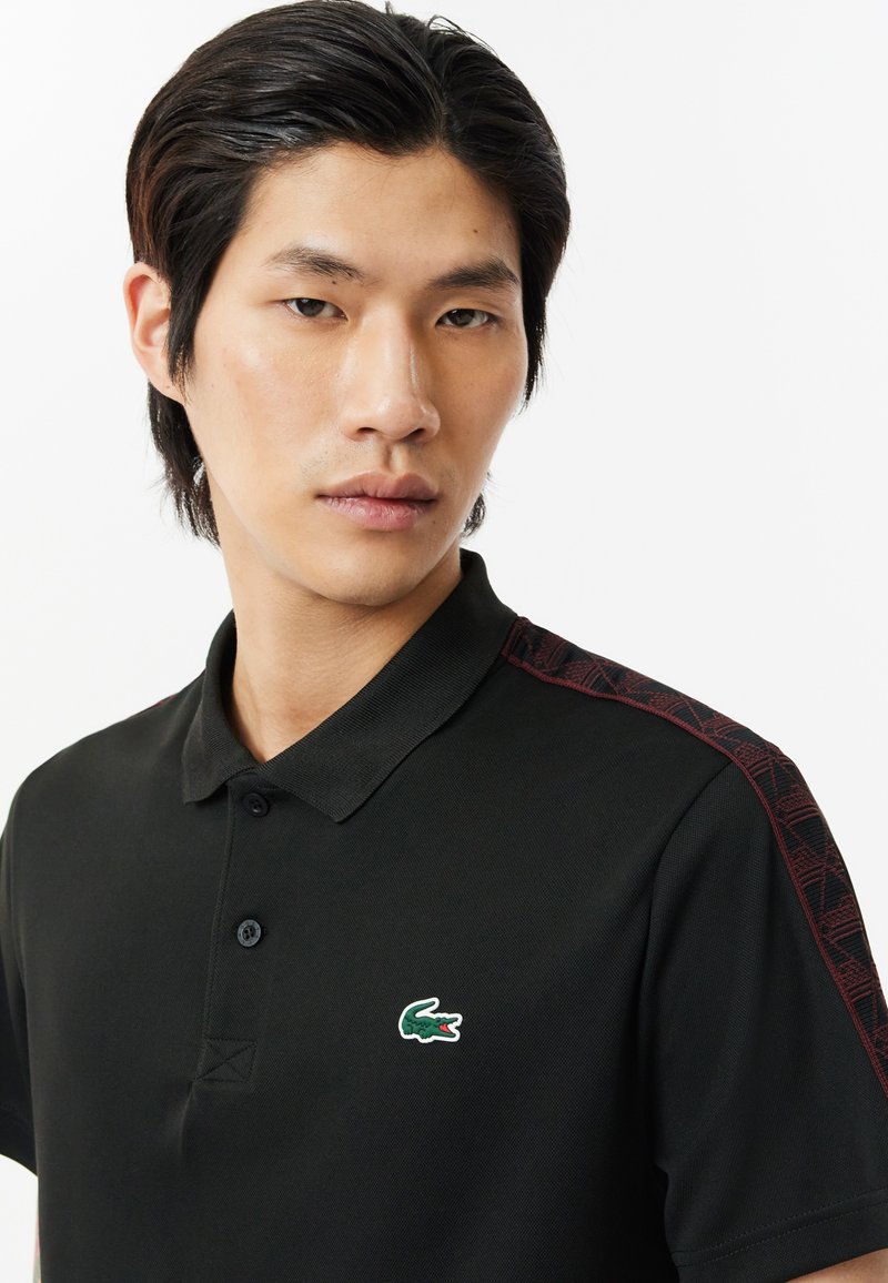 Schwarzes Polohemd mit einem strukturierten roten Musterdesign an den Schultern. Verfügt über einen knöpfbaren Kragen und ein grünes Krokodil-Logo auf der Brust.