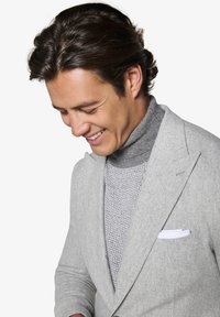 Lys grå blazer i en tekstureret uldblanding, båret over en grå strik turtle neck. Hvid lommefyrkant synlig, hvilket skaber et lagdelt look.