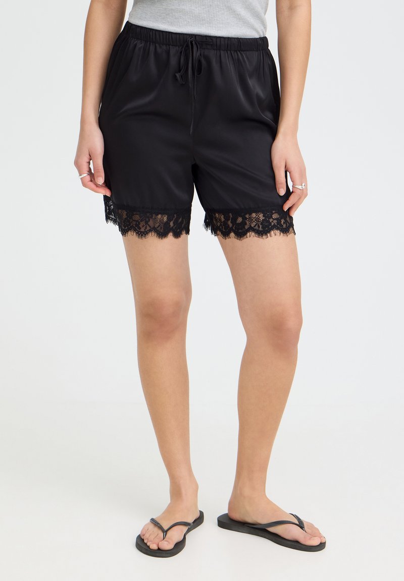 Femme portant un short en satin noir avec bordure en dentelle, un débardeur gris, des bagues en argent et des tongs noires, debout devant un fond uni.