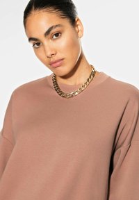 Overdreven lichtbruine sweatshirt met een ronde halslijn en korte mouwen, gecombineerd met een flinke gouden ketting. Soepele textuur.