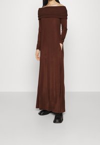 Lång, brun av-axeln-klänning med långa ärmar, gjord av ett mjukt material. Har sidofickor och en avslappnad, flödande silhuett.