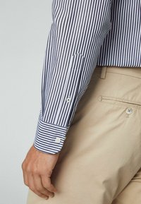 Uomo che indossa una camicia a righe verticali blu e bianche con polsini abbottonati e pantaloni beige con tasca posteriore abbottonata.