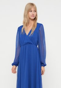 Mulher loira vestindo um longo vestido azul com mangas longas transparentes, olhando ligeiramente para o lado contra um fundo branco simples.