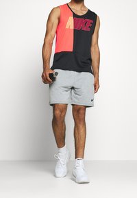 Débardeur Nike à blocs de couleur en rose vif et noir, arborant un grand logo. Associé à un short gris et des baskets blanches. Tenant une bouteille d'eau.