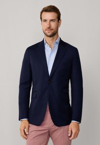 Blazer blu navy con taglio su misura, revers a scudo, due tasche frontali e due bottoni. Indossato sopra una camicia azzurra e pantaloni rosa.