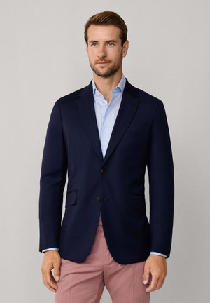 Blazer blu navy con taglio su misura, revers a scudo, due tasche frontali e due bottoni. Indossato sopra una camicia azzurra e pantaloni rosa.