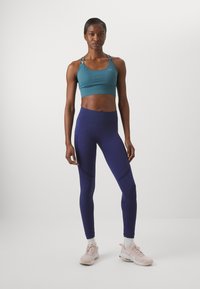 Sujeción deportiva de color azul verdoso con relieve, leggings púrpuras de cintura alta y zapatillas deportivas de color rosa claro. Los leggings presentan detalles de diseño sutiles y una textura suave.