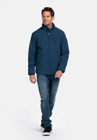 Blauwe softshell jas met een hoge kraag, een rits aan de voorkant en een borstzak; gecombineerd met denim jeans en casual schoenen.