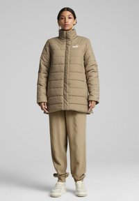 Khaki värvi puffjakker kõrge kraega ja tikandiga disainiga. Vasakul rinnal on Puma logo. Kombineeritud beežide tekstureeritud pükstega ja tossudega.