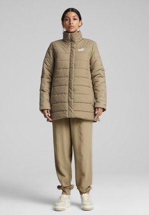 Khaki puffer bunda s vysokým golierom a prešívaným dizajnom. Obsahuje logo Puma na ľavej hrudi. Skombinovaná s béžovými textúrovanými nohavicami a teniskami.