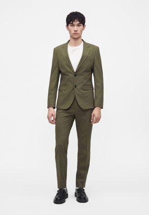 Uomo che indossa un completo verde oliva con scarpe nere e una maglietta bianca con scollo rotondo, in piedi davanti a uno sfondo bianco uniforme.