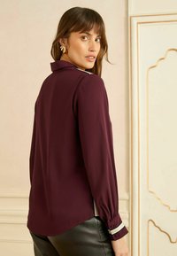 Burgundia pikkade varrukatega pluus kraega, mansetil nööpdetailidega ja kergelt läikiva pinnaga. Sobitatud mustade kitsaste pükstega.