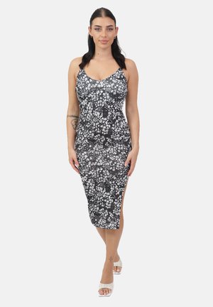 Vrouw draagt een zwart-witte midi-jurk met bloemmotief, spaghettibandjes en een zijsplit, gecombineerd met witte open-toe hakken, staand tegen een witte achtergrond.