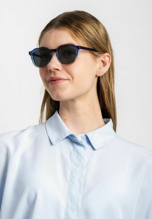 Blå-gradient solbriller med en tyk, rund ramme. Grå linser. Båret med en lyseblå skjorte med krave. Glat tekstur.
