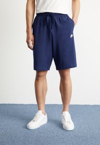 Mornarško modri atletski šortsi z elastičnim pasom in belim logotipom Nike na levi strani, v kombinaciji z belimi supergami, zasnovani za udobje.