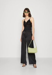 Moteris dėvi juodą nėriniuotą kamisolę, plačiomis satino kelnėmis, žalius akinius nuo saulės, laiko šviesiai žalią rankinę ir juodus basutus su dirželiais, fone baltas fonas.