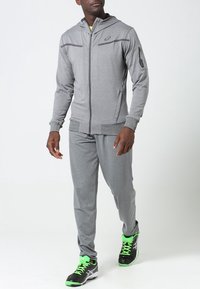 Šedý sportovní teplákový komplet s kapucí, se zipem vpředu, bočními kapsami a černými akcenty. Nosí se s černými a zelenými teniskami.