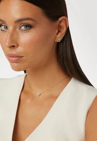 Goldene, herzförmige Ohrringe und ein passender Anhänger an einer feinen Kette mit klaren Edelsteinelementen, getragen von einem Modell vor einem hellen Hintergrund.