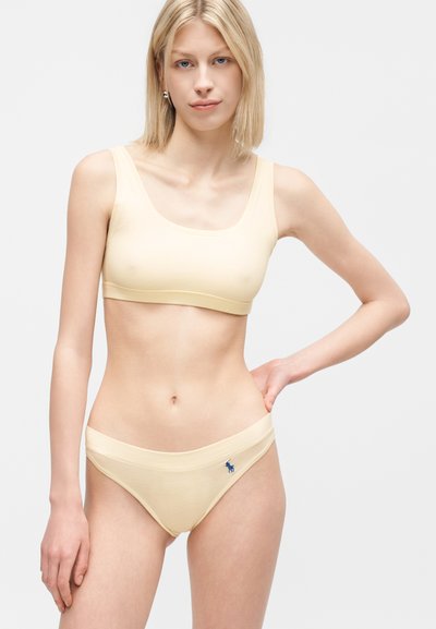 Νεαρή γυναίκα που φοράει μπεζ αθλητικό μπουστάκι και αντίστοιχο εσώρουχο με μικρό ναυτικό λογότυπο, στέκεται μπροστά σε απλό ανοιχτό γκρι φόντο.