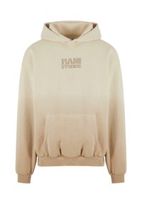 Beige oversized hoodie med en framficka, muddar vid ärmarna och logotypen "KANI STUDIOS" i tonad tryck på bröstet. Slät tygtextur.
