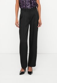 Čierne nohavice s priliehavým strihom, výraznými záhybmi, vysokým pásom a hladkou textúrou, v kombinácii so špicatými čiernymi lodičkami.