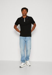 Camiseta polo negra con cuello a rayas, combinada con jeans azul claro y zapatillas blancas. El modelo está de pie con las manos en las caderas. Fondo liso.