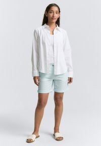 Chemise blanche à manches longues avec des boutons sur un débardeur bleu clair, accompagnée de shorts menthe à rouleaux et de sandales à glissière beige.