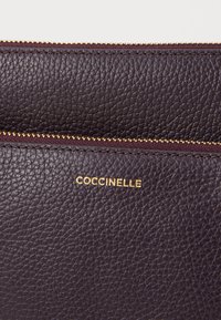 Pochette in pelle viola scuro con superficie testurizzata, chiusura con zip dorata e logo "COCCINELLE" in rilievo dorato sul davanti.