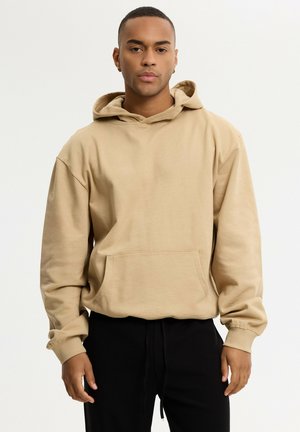 Joven con sudadera con capucha beige con bolsillo delantero y pantalones negros, de pie frente a un fondo claro y liso.