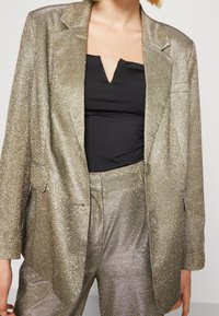 Blazer dorado metálico con una superficie texturizada, con solapas, dos bolsillos frontales y combinado con pantalones a juego.