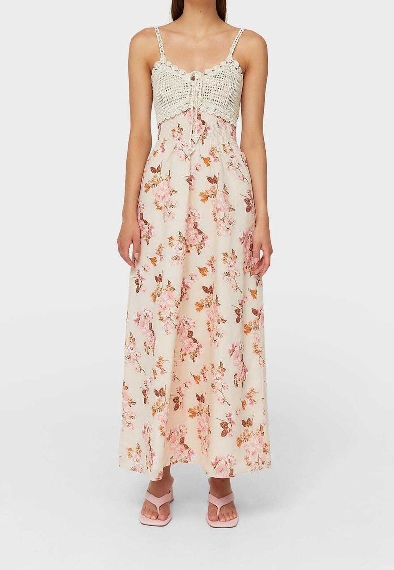 Femme portant une longue robe crème avec un haut en crochet, des bretelles fines et une jupe à fleurs roses, debout en sandales rose clair sur un fond uni.