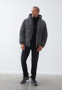 Graue Daunenjacke mit Kapuze, gestepptem Muster, Frontreißverschluss und zwei Seitentaschen. Kombiniert mit einem dunklen Rollkragenpullover und schwarzen Jeans, dazu Sneaker.