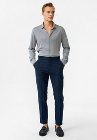 Lichtgrijs overhemd met lange mouwen, marineblauwe op maat gemaakte broek en donkere loafers. De outfit presenteert een strakke, gestroomlijnde uitstraling.