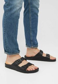 Svarta gummisandaler med två justerbara spännremmar, bärs med upprullade blå denimjeans, med en texturerad sula.