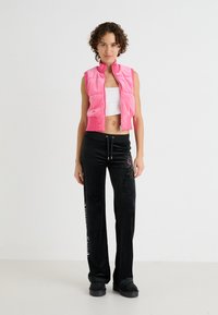 Vesta puf roz, fără mâneci, cu fermoar frontal, asortată cu pantaloni negri, largi, ce prezintă accente decorative și un brâu cu șnur.