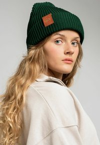 Zielona, prążkowana czapka beanie ze skórzaną metką w kolorze brązowym, noszona z beżowym, wysokim swetrem z kołnierzem. Długie, faliste włosy częściowo widoczne.