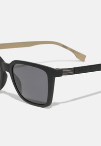 Sorte rektangulære solbriller med mørke linser og behandlet overflate. Stengene har en kontrasterende lys tannfarge med metalliske detaljer.