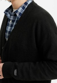Černý cardigan s jemnou texturou, zapínáním na knoflíky a ribbovanými manžetami, navlečený přes modro-bílou kostkovanou košili.