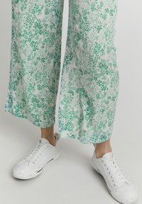 Pantalones holgados con patrón floral en tela ligera, presentando tonos verdes sobre un fondo blanco, combinados con zapatillas blancas.