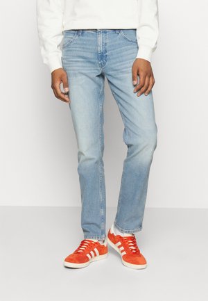 Homme portant un jean bleu clair, un sweat-shirt blanc, des chaussettes blanches et des baskets orange vif avec des rayures blanches, se tenant devant un fond uni.