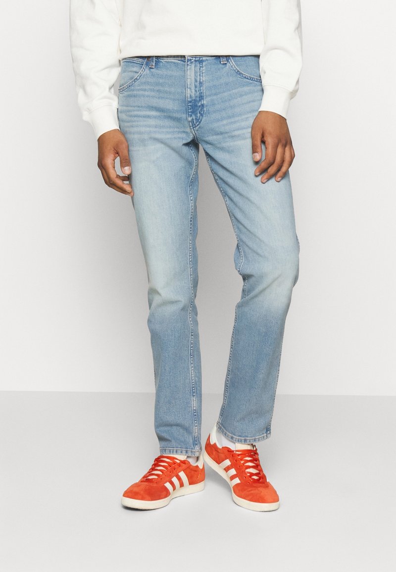 Uomo che indossa jeans azzurri chiari, felpa bianca, calzini bianchi e scarpe da ginnastica arancioni vivaci con strisce bianche, in piedi contro uno sfondo semplice.