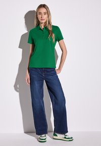 Grön kortärmad polo, avslappnade blå jeans och gröna och vita sneakers. Enkel design med knappslå, avslappnad passform.