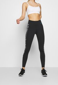 Černé legíny s vysokým pasem, s krajkovaným bočním panelem. Přiřazené s bílým crop topem a černými sportovními botami.