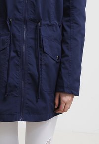 Veste à capuche bleu foncé avec fermeture éclair, taille ajustable à cordon, deux poches avant, et un tissu lisse et texturé.
