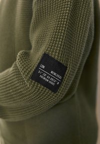 Grønt strikket stof med sort rektangulært mærke, der viser "LDN," romertal, koordinater og teksten "Premium Goods."